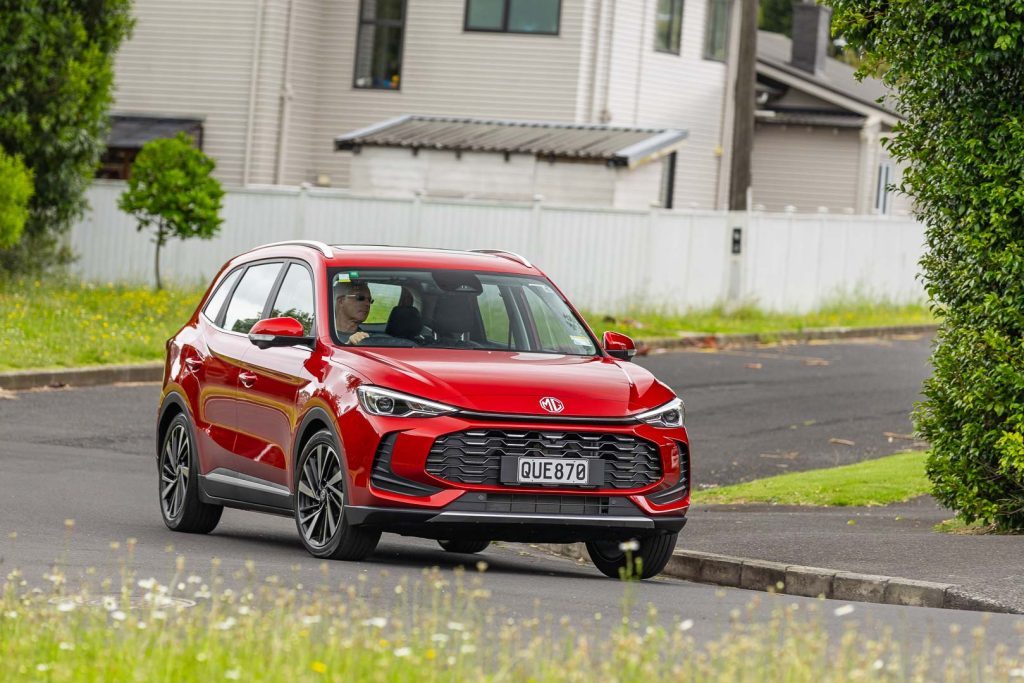 Cornering shot of a red 2024 MG ZS Hybrid+ Essence, with wild flowers in the foreground