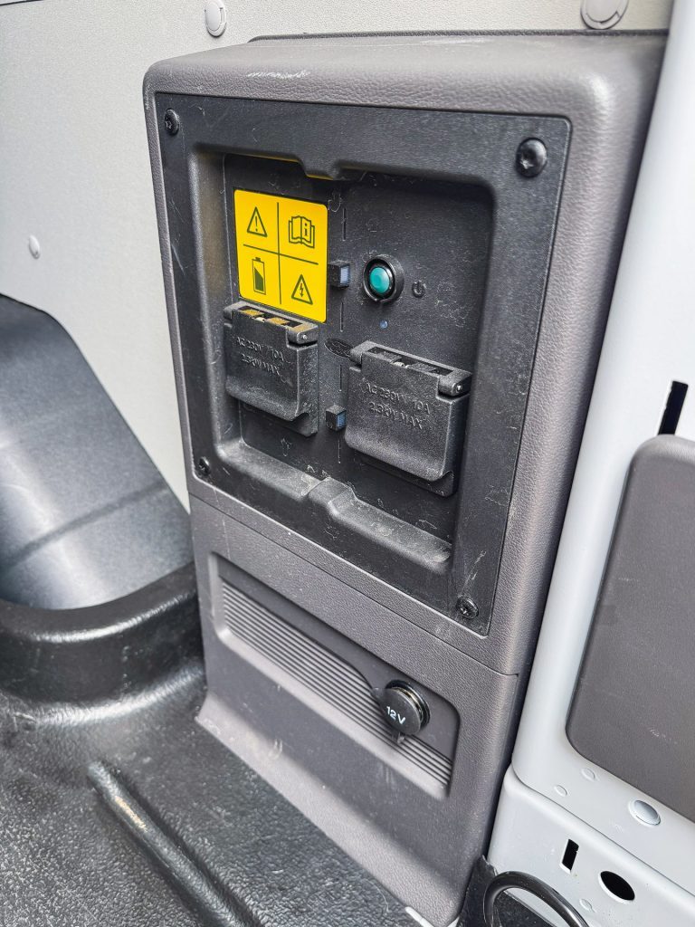 A vehicle's interior panel with two covered AC outlets, a 12V outlet, and yellow warning labels