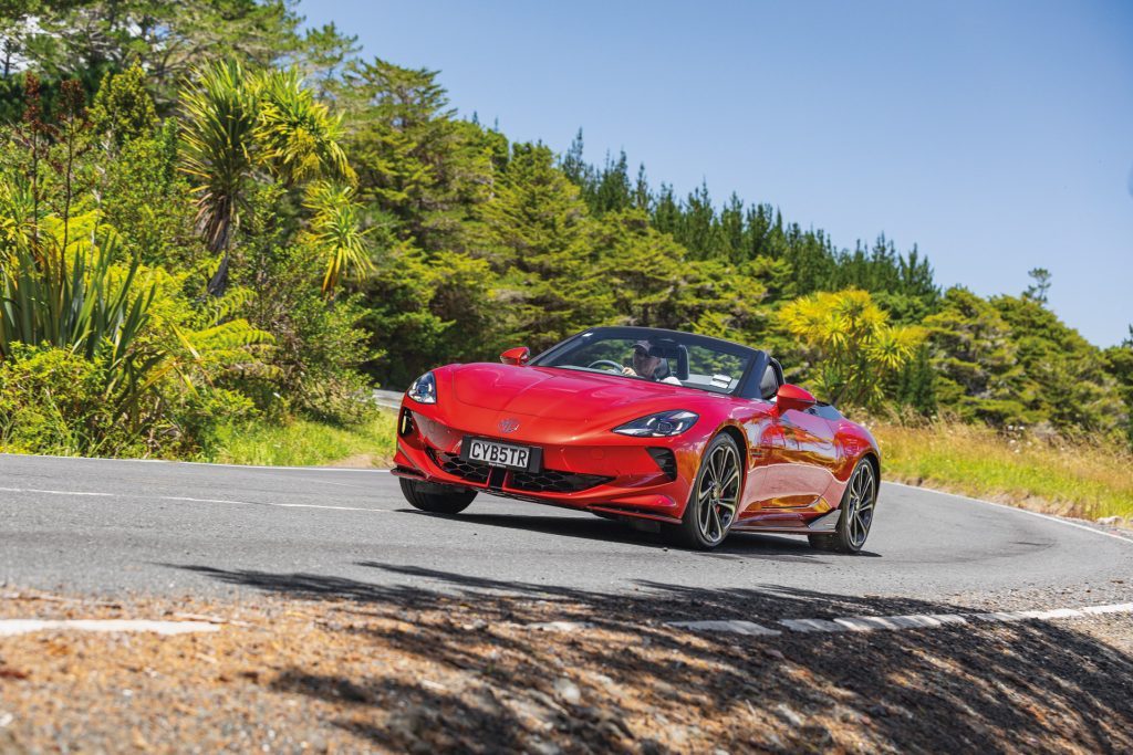 Red MG Cyberster convertible with scissor doors and dual-motor system, driven by a man on a scenic road