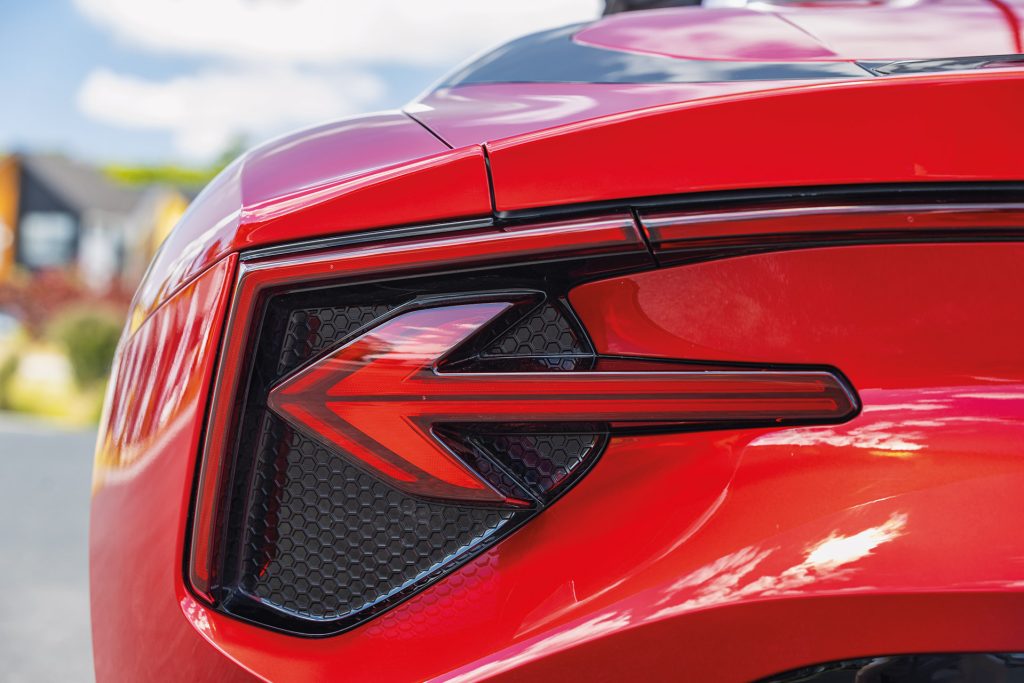 "Close-up of the rear left taillight of a red sports car with a distinctive arrow-shaped design and red and black accents, reflecting the surroundings