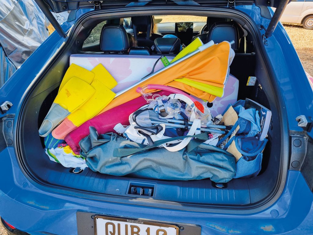 Open trunk of a blue Ford Mustang Mach-E electric SUV filled with various items for a beach outing, showcasing its practical storage capacity