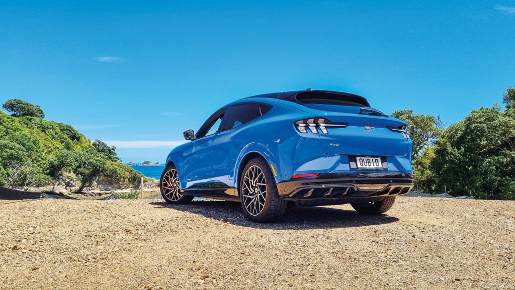 A blue Ford Mustang Mach-E electric SUV parked on a gravel surface with a scenic background of greenery and a clear blue sky