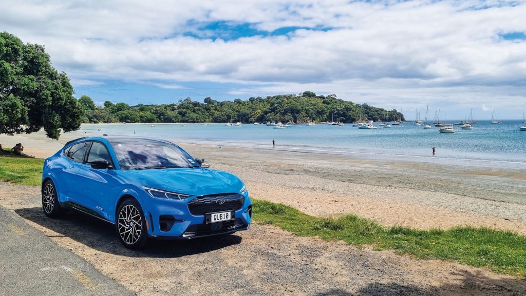 A blue Ford Mustang Mach-E electric SUV parked near a beach with calm waters, boats, and tree-covered hills in the background