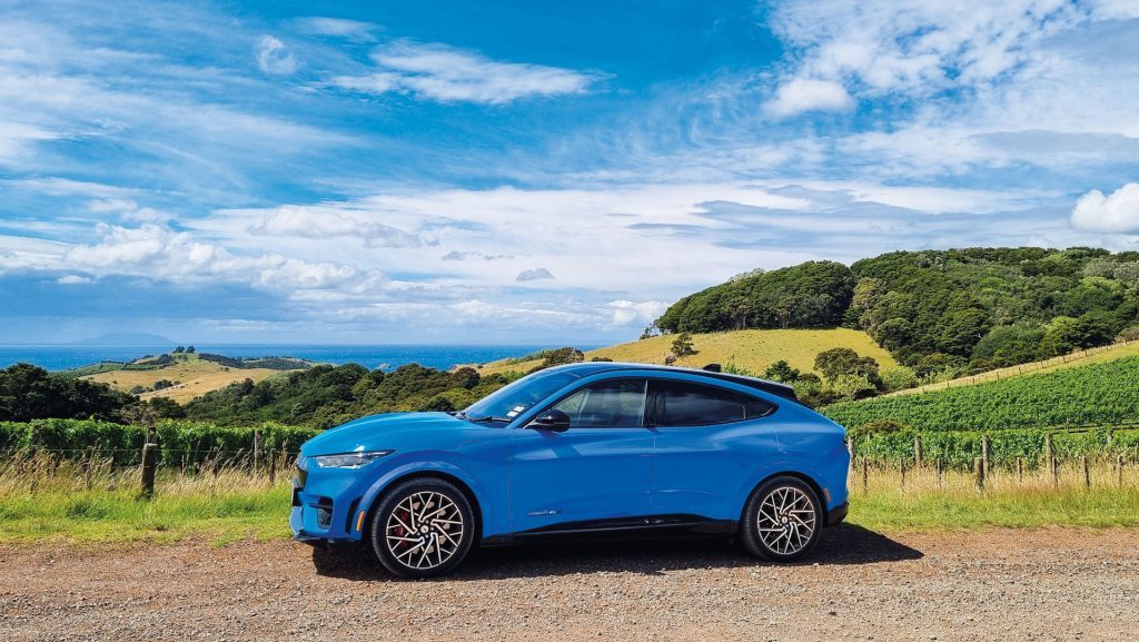 A blue Ford Mustang Mach-E electric SUV parked on a scenic countryside gravel road, with rolling green hills, a fence, and a distant ocean view under a partly cloudy sky