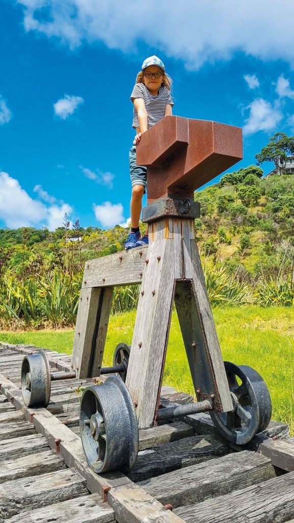 A large sculpture named Rua-Kuriwao, showcasing a rusted metal structure mounted on a wooden rail cart with metal wheels, positioned on wooden tracks. The background features a grassy area with bushes, trees, and a clear blue sky with a few clouds