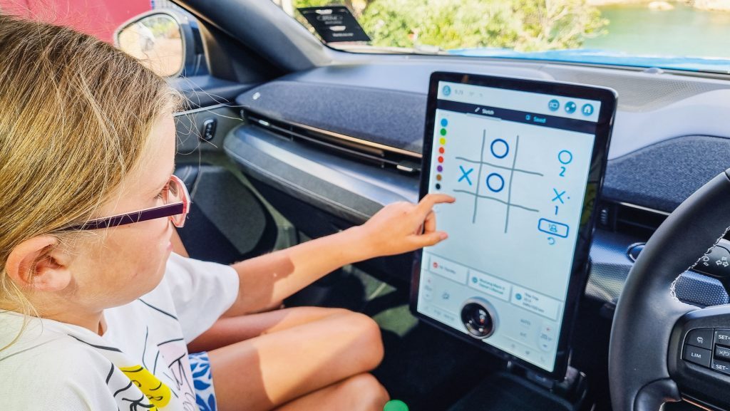 A child sitting in the front passenger seat of a blue Ford Mustang Mach-E playing tic-tac-toe on the vehicle's touchscreen display while waiting for the ferry ride to Waiheke Island