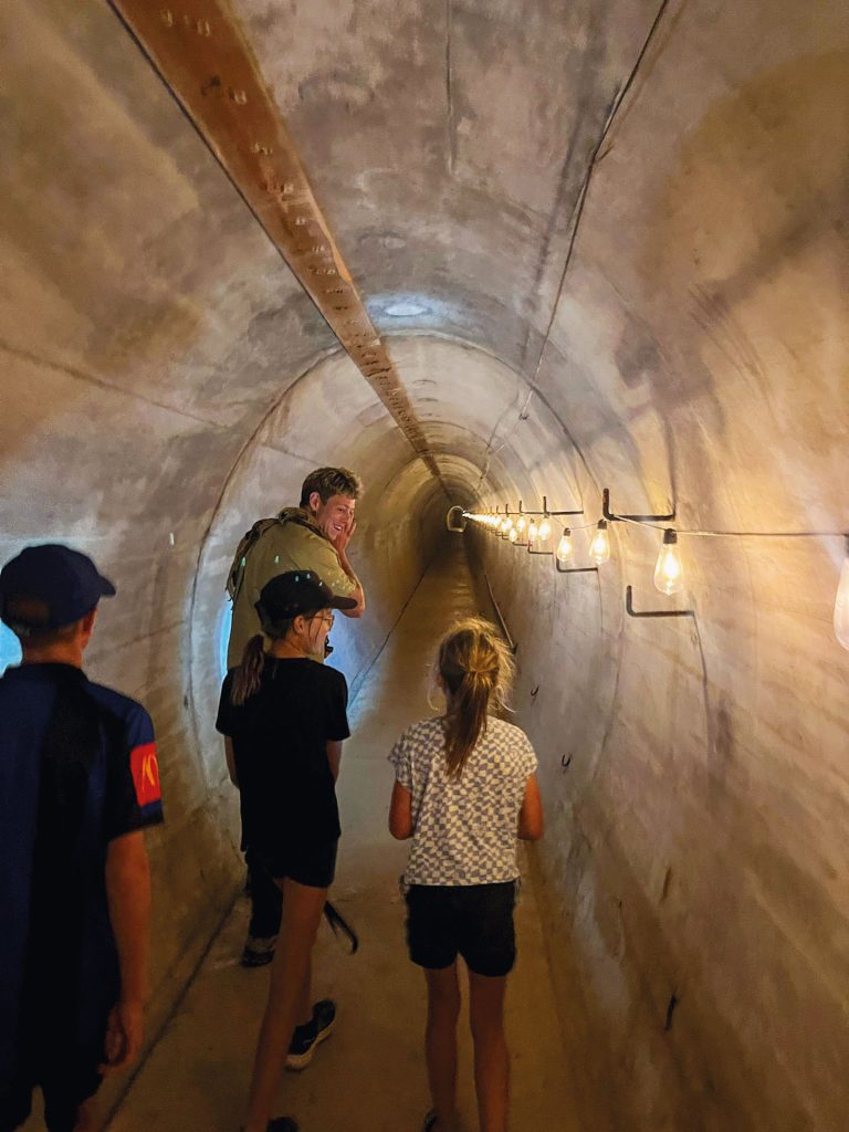 A family exploring an underground tunnel with concrete walls, illuminated by a series of exposed light bulbs on the right side