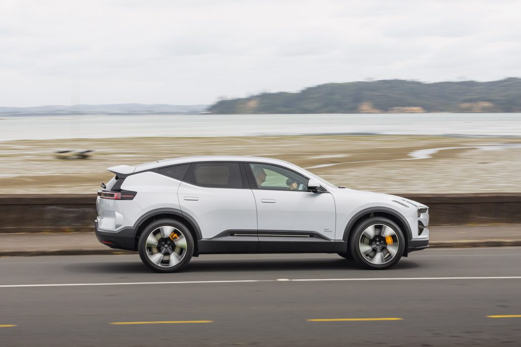 A white Polestar 3 electric SUV driving along a coastal road, showcasing its sleek design and scenic waterfront background